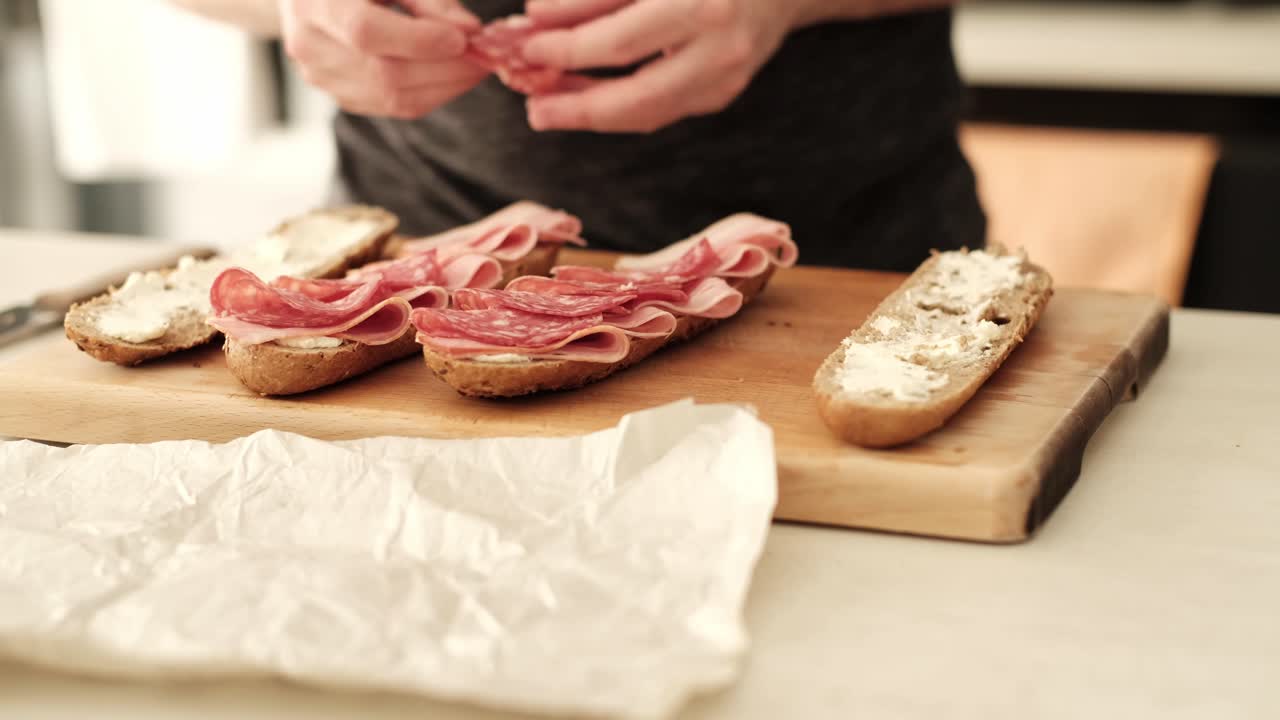 Caucasian male placing Italian salami on a halved brown baguette in the kitchen on the countertop