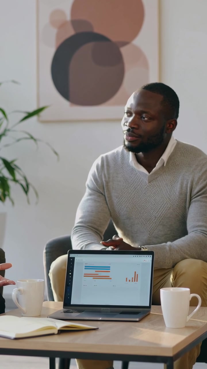 Vertical video: Listening man in gray sweater reviewing charts on laptop in office, peer gesturing