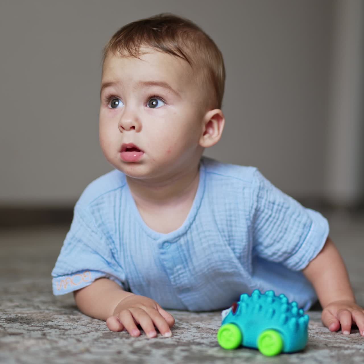 Lovely baby boy in blue clothes lies on his belly on the floor. Cute child has a toy in front of him but looks around with interest