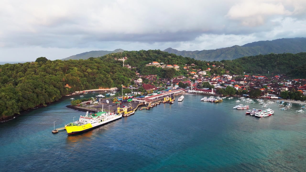 Porto di Padang Bai a Bali con un traghetto passeggeri attraccato al molo, sullo sfondo di lussureggianti colline verdi e una baia turchese con barche ancorate, suggestive riprese aeree con drone.