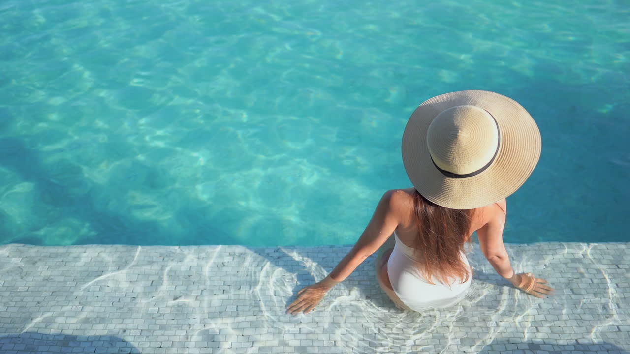 Glamour Female in Swimsuit and Summer Hat Sitting on Swimming Pool Edge on Sunny Day