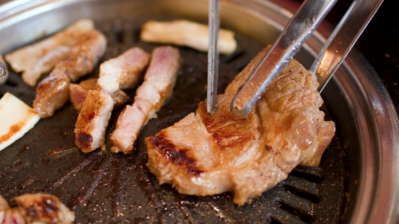 Hand uses metal tongs to flip grilled pork on round Korean barbecue grill, warm lighting