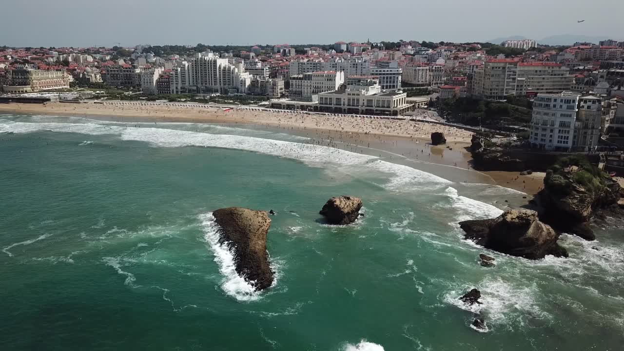 Costa de Biarritz, la famosa Grande Plage, playa llena de gente y olas rompiendo en icónicos farallones, País Vasco, Francia. Vista panorámica aérea con dron