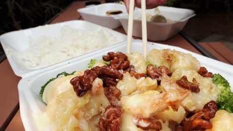 Close-up-shot-of-person-eating-takeout-Singaporean-Honey-Walnut-Shrimp-with-rice-and-broccoli-using-chopsticks