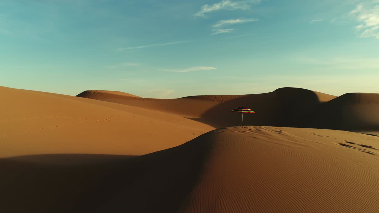 Aerial fly by of colorful beach umbrella isolated in desert sand dunes at sunset