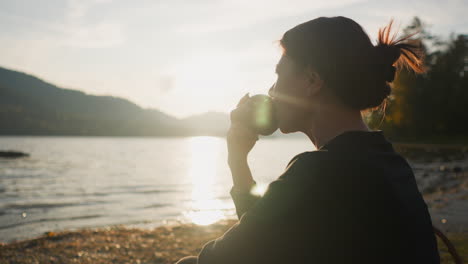 lonely lady eats apple on river bank at sunset. sad woman eats fruit alone with nature. incredible view of lake and rolling mountains in sunny autumn weather