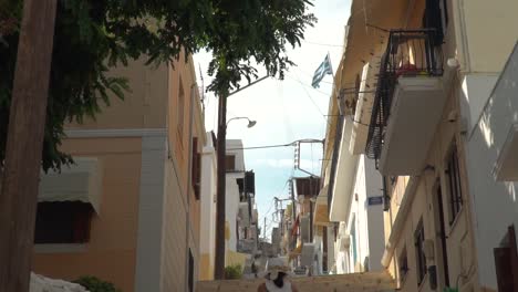 Tilt-up-from-girl-in-summer-dress-to-Greek-flag-walking-up-steps-in-narrow-alley-in-Syros