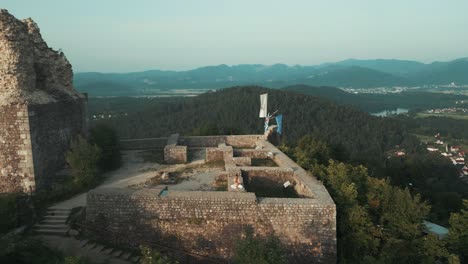 Tiro-De-Dron-De-Un-Hombre-Indio-Sentado-En-Pose-De-Hatha-Yoga-Y-Meditando-Nadhi-Shuddhi-En-La-Pared-Del-Castillo-De-Piedra-En-La-Cima-De-La-Colina-Rodeada-De-Colinas,-Campos,-Lago-Y-Bosques-En-La-Mañana-Al-Amanecer