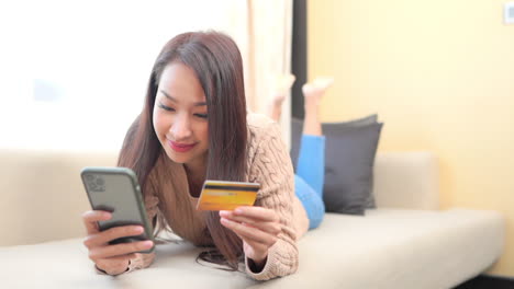 young smiling asian girl lying on sofa holding credit card and smartphone