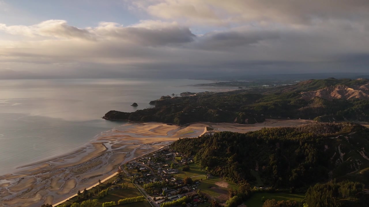 Cidade de Marahau aninhada entre colinas arborizadas e planícies de maré da Baía de Tasman, com a luz dourada do nascer do sol rompendo as nuvens de tempestade sobre o estuário e promontórios distantes. Nova Zelândia. Tomada aérea ampla.