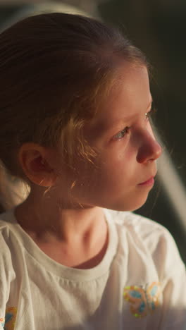 sunlit child looks at sunrise in room. thoughtful little girl looks at nature outside window. pretty kid rests sitting in soft chair on terrace