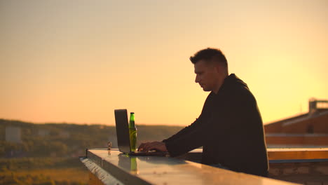 a male freelance programmer sits on a skyscraper roof with a laptop and beer typing code on a keyboard during sunset. remote work.