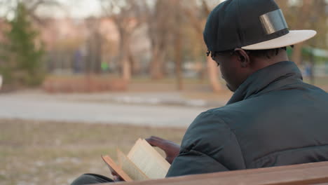 rear view of young man wearing cap and winter jacket sitting on park bench, flipping through pages of open book with focus, blurred outdoor background creating calm and reflective atmosphere