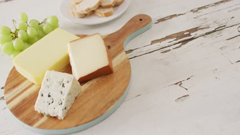 video of cheeses on chopping board, with grapes and bread on rustic wooden table with copy space