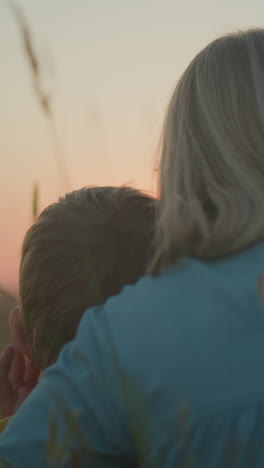 vista de fondo de una madre en un vestido azul sentada en un campo con sus dos hijos pequeños a su lado. todos están viendo la serena puesta de sol, con la luz cálida proyectando un resplandor pacífico en la escena