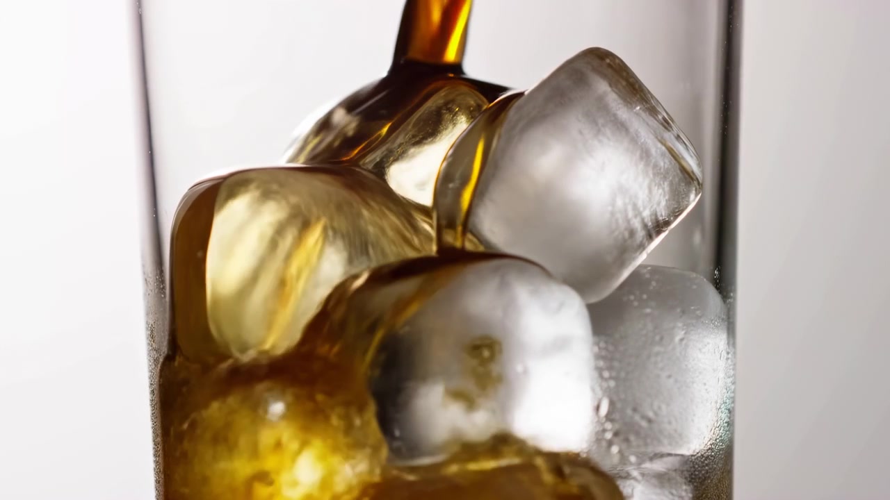 Primo piano di una bevanda ambrata che scorre in un bicchiere trasparente pieno di cubetti di ghiaccio grandi, con una retroilluminazione brillante e uno sfondo pulito e minimalista. Il liquido.
