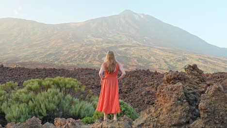 girl in orange summer dress standing on lava rocks, looking at volcano teide