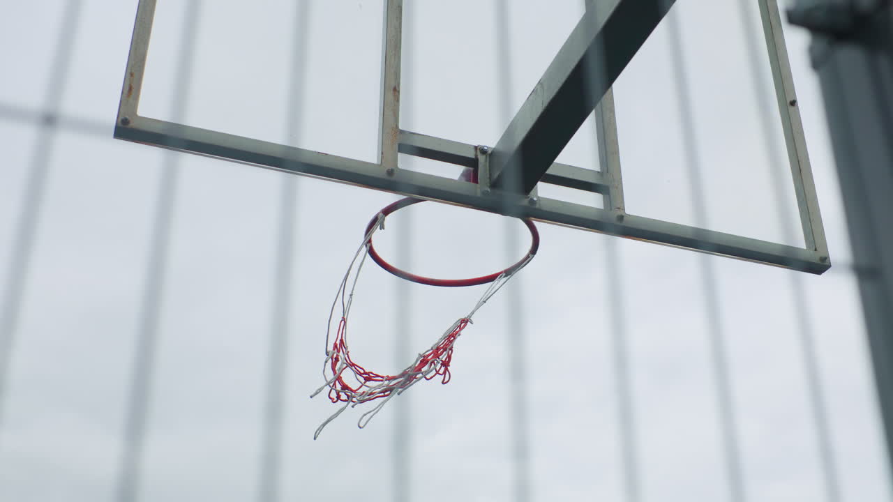 primo piano di un canestro da basket con rete increspata, palla assente, rete che si attorciglia verso il basso dopo un netto canestro, bordo metallico luccicante, supporto del tabellone visibile, luce nuvolosa intensa, dettagli ad alta velocità dell'otturatore, esterno