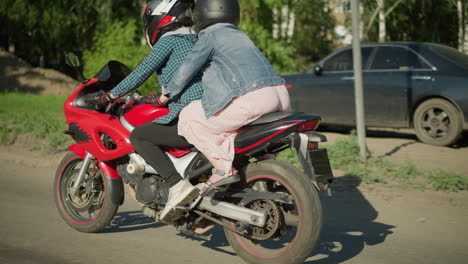 two women riding on a power bike along an urban road, the passenger holds onto the rider as they travel past trees and buildings