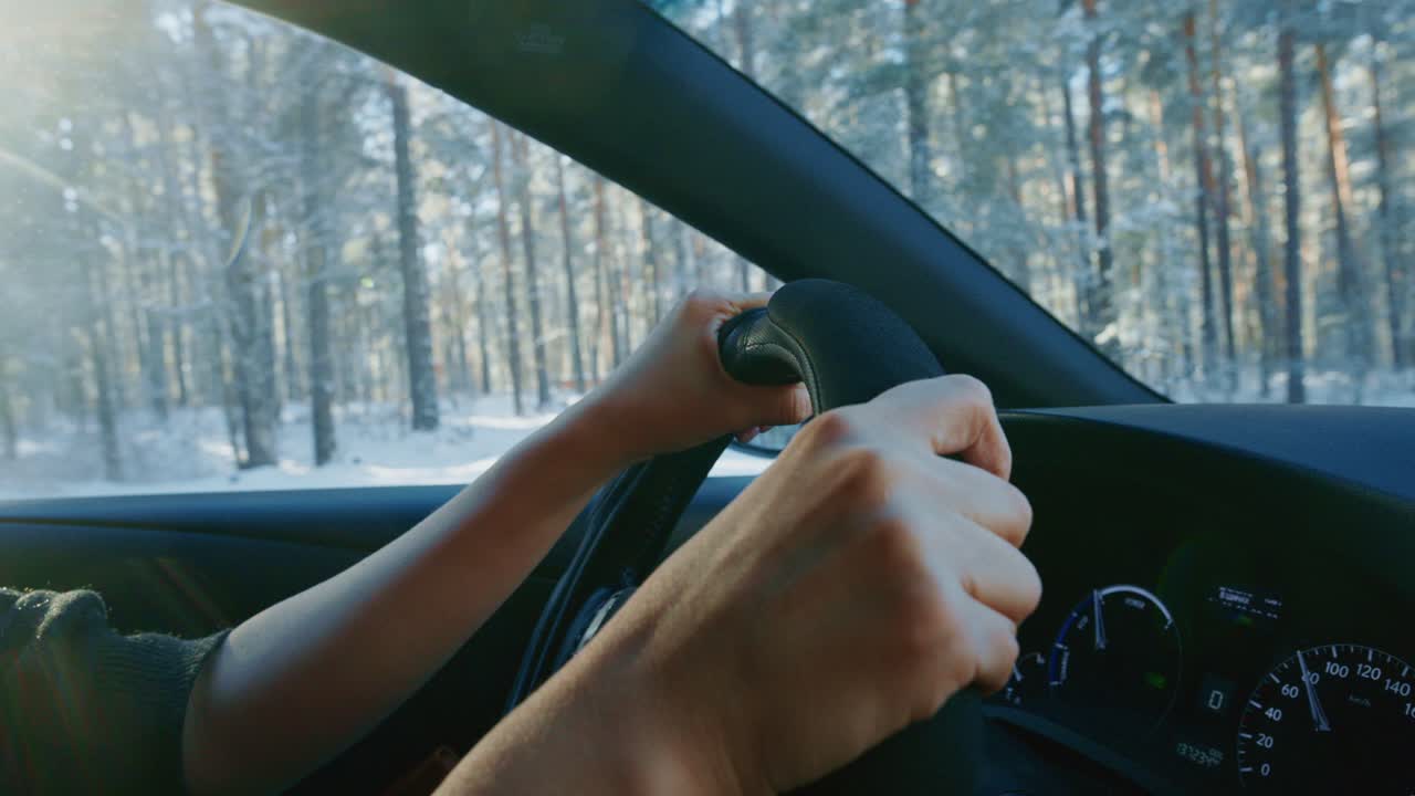 Hænder griber fat i rattet, mens bilen finder vej gennem en sneklædt skov. Det fredfyldte vinterlandskab er fyldt med høje træer, der skaber en rolig atmosfære på køreturen.