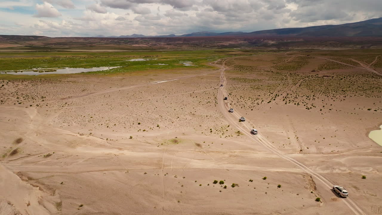 Tourist convoy of 4x4 vehicles on road trip through Siloli Desert, adventure travel in Bolivian Altiplano landscape with oasis, Bolivia. Aerial drone orbiting