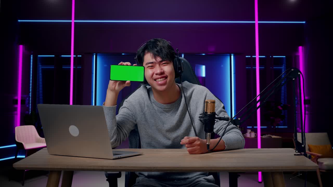 A Smiling Asian Man With Headphone And Computer Showing Green Screen Mobile Phone In Room Lit By Neon Lights In Retro