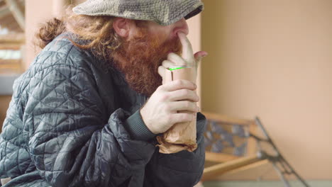 hombre sin hogar al aire libre cerca de un banco roto hurgándose la nariz con lata de cerveza en las manos