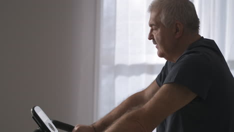 sport-activity-during-self-isolation-man-is-training-with-stationary-bicycle-at-home-portrait-of-grey-haired-person-portrait-shot