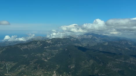 an aerial view of tramuntana range (tramontana), in the north of mallorca island, taken from a plane cockpit, with some cottony clouds under a bright blue sky.