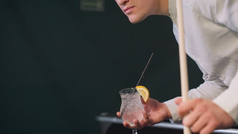 close-up of hand holding cue stick and lemon drink while pacing in billiards hall. ice cubes and lemon slice in glass create a refreshing contrast against dimly lit background with green pool table