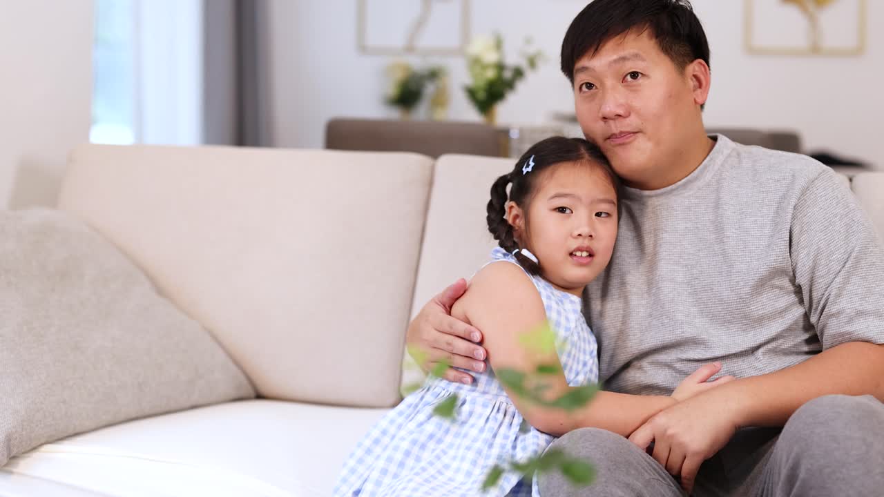 Asian father comforts scared daughter on modern sofa, reacting to frightening TV scene at home