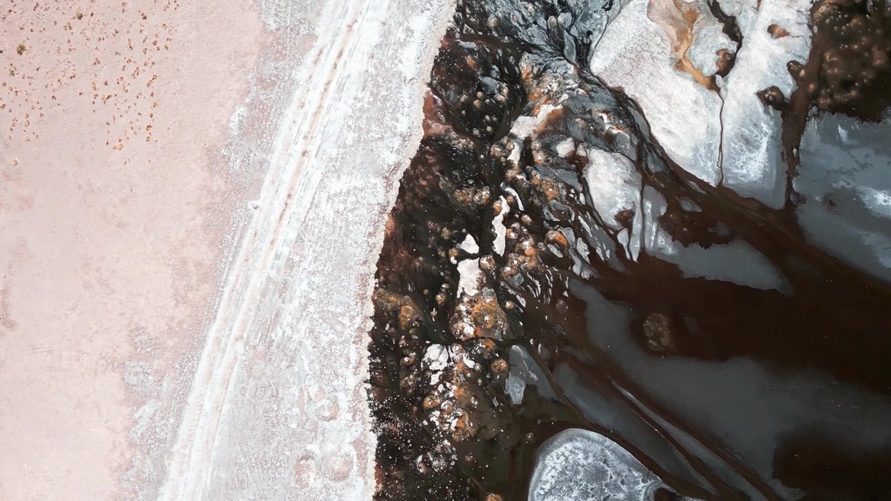 Vista aerea dall'alto della costa della Laguna Tara con crosta di sale rosa, depositi minerali scuri, sedimento arancione e acqua scura e immobile che creano vivaci motivi astratti. Laguna Tara, San Pedro de Atacama, Cile