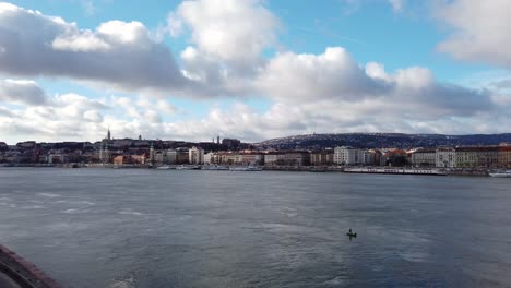 Toma-Panorámica-Sobre-El-Río-Danubio-En-Budapest,-Hungría-Con-Las-Colinas-De-Buda-Y-El-Parlamento-Al-Fondo,-El-Edificio-De-Oficinas-Del-Parlamento-En-Primer-Plano,-Tomada-Desde-El-Puente-Margaret