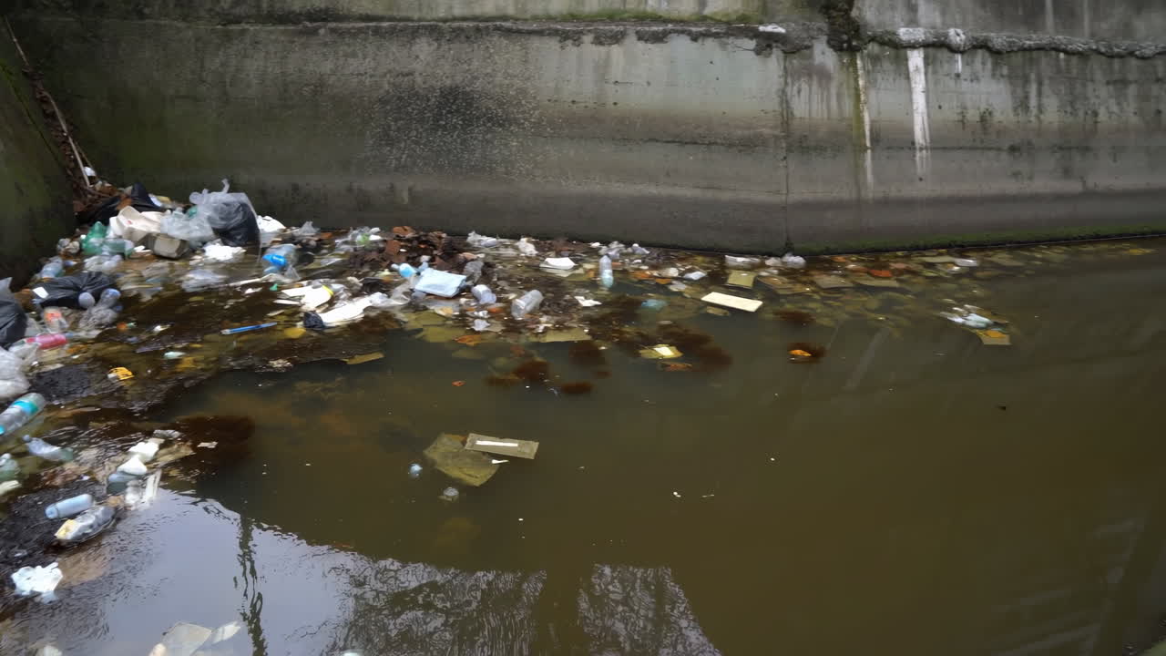 Canal Water Clogged With Plastic Bottles And Bags Against Concrete Wall, Murky Brown Surface With Floating