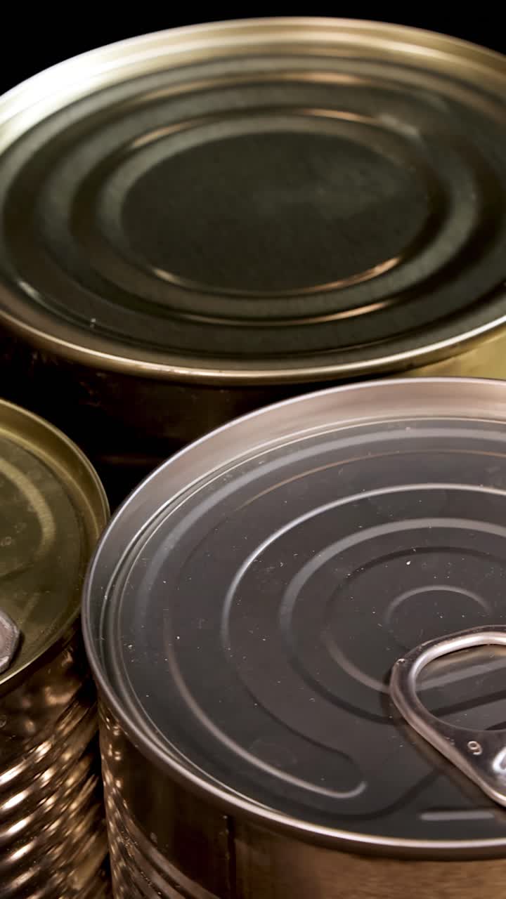 Latas de alimentos metálicas de varios tamaños giran bajo una iluminación de estudio cálida, destacando las pestañas de apertura y las texturas. El movimiento de la cámara es suave, manteniendo un primer plano.