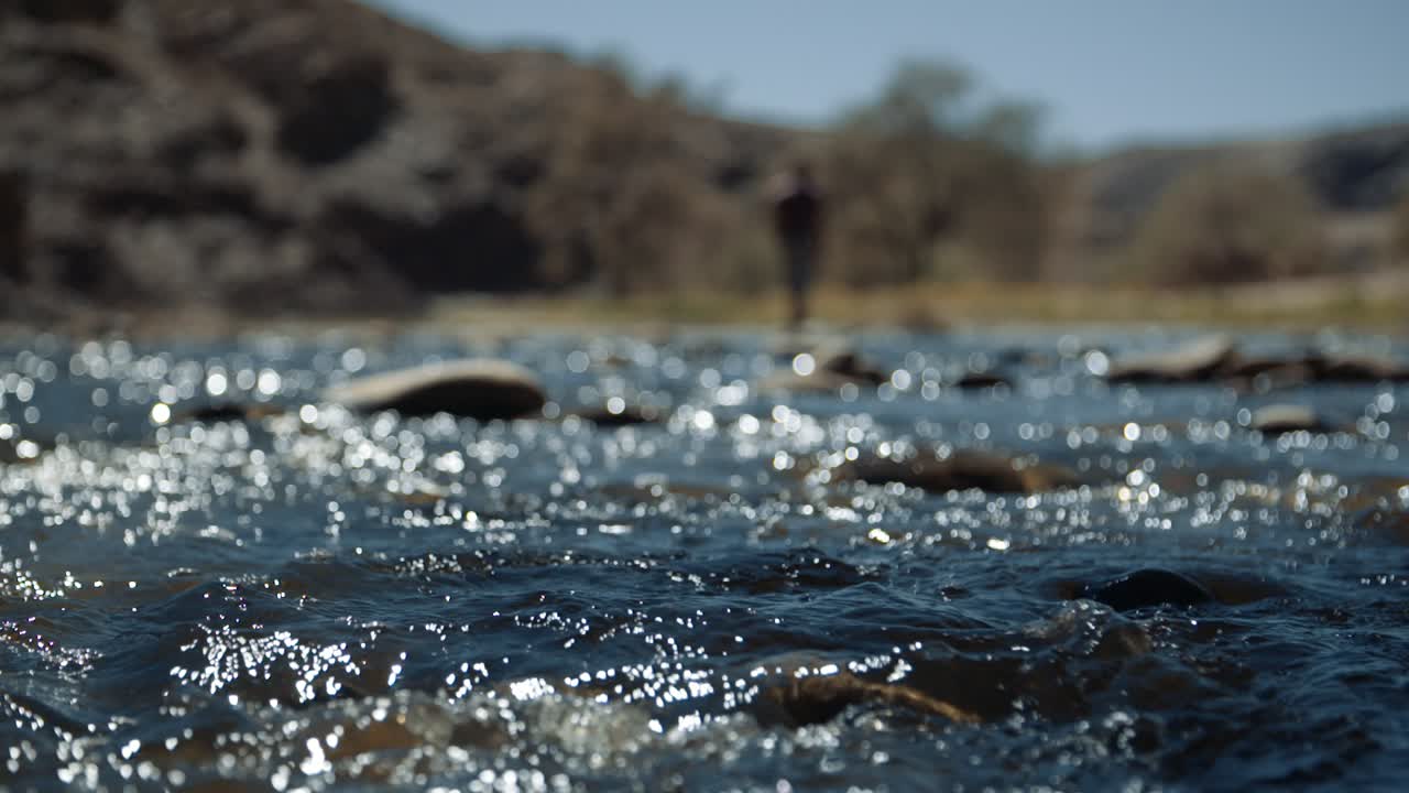 Ripresa a mano libera in slow motion con profondità di campo ridotta di un fiume che scorre in Africa mentre un uomo cammina sullo sfondo.