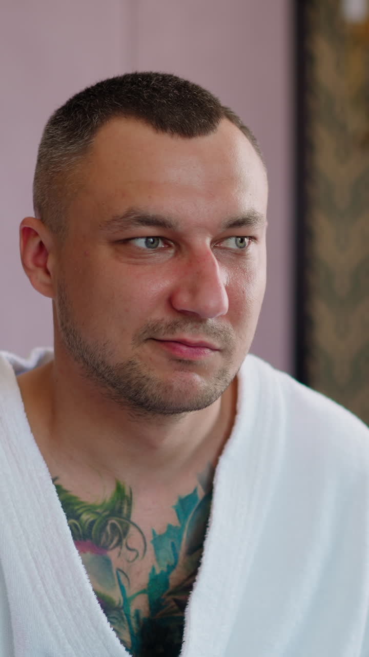 Angry man with three-day stubble and colorful tattoos on chest in bathrobe grimaces sitting on bed in comfortable hotel room closeup slow motion