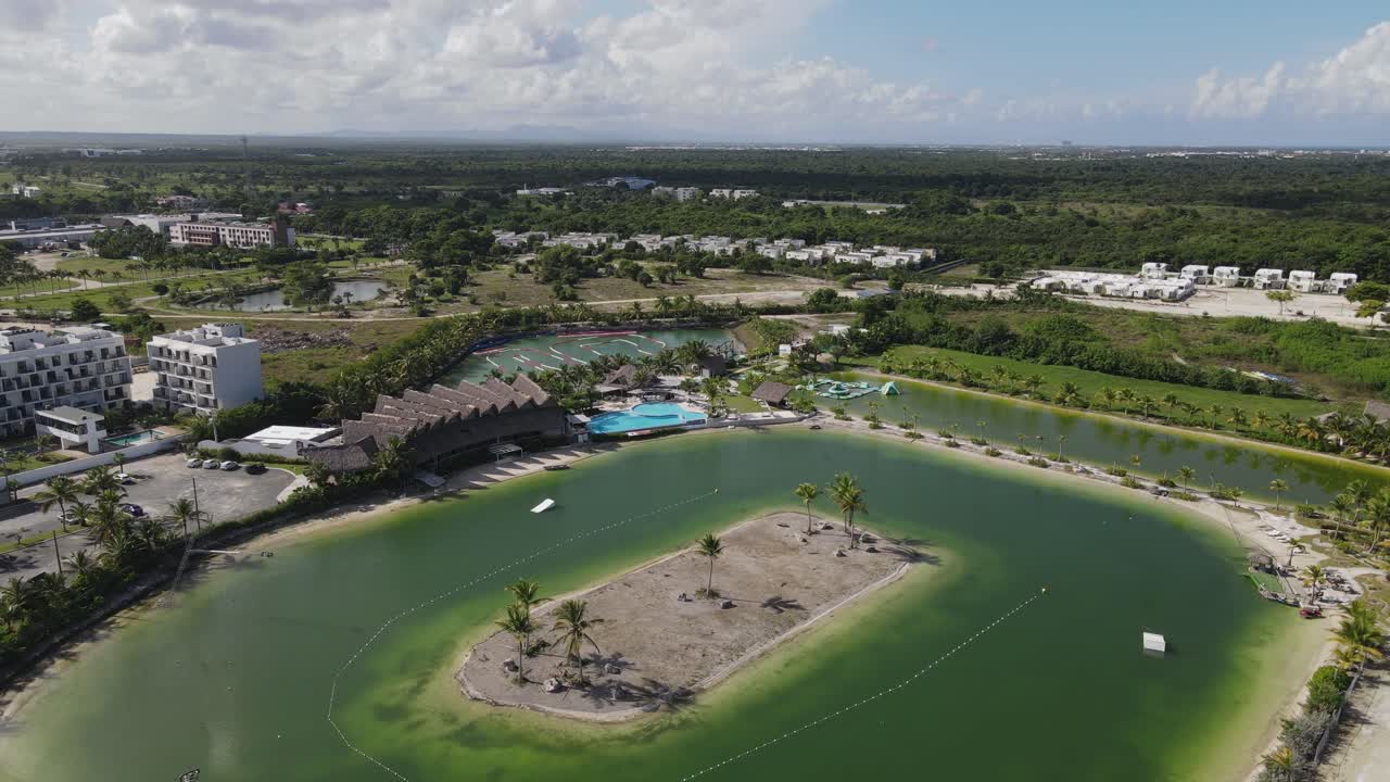 Caribbean Lake Park, featuring lush greenery and a winding lake. The park includes sandy areas, palm trees, and recreational spaces, ideal for relaxation and outdoor activities. Aerial wide shot.