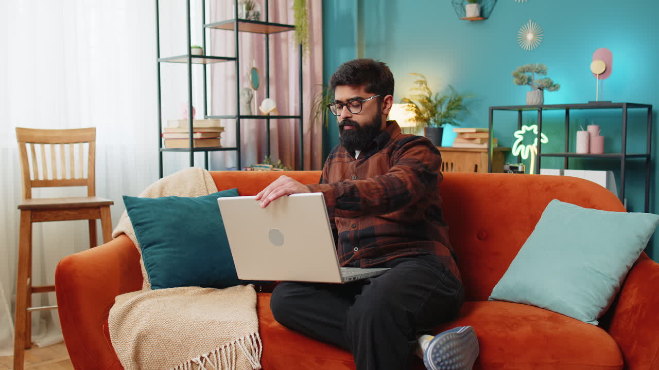 Happy young indian man typing working on laptop browsing internet using apps sitting on home sofa