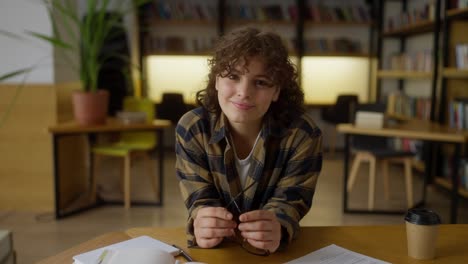 ritratto di una felice studentessa con i capelli ricci in una camicia a quadri che posa mentre è seduta al tavolo e tiene gli occhiali in mano nella biblioteca