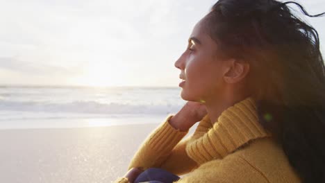 Portrait-of-thoughtful-hispanic-woman-sitting-on-beach-at-sunset
