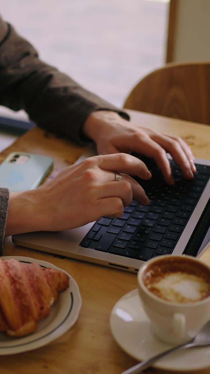 Mani che digitano sulla tastiera del laptop con caffè e croissant al bar