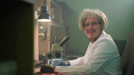 una vista de cerca de un técnico sonriente con una peluca rubia y gafas, trabajando en un escritorio de laboratorio con un microscopio