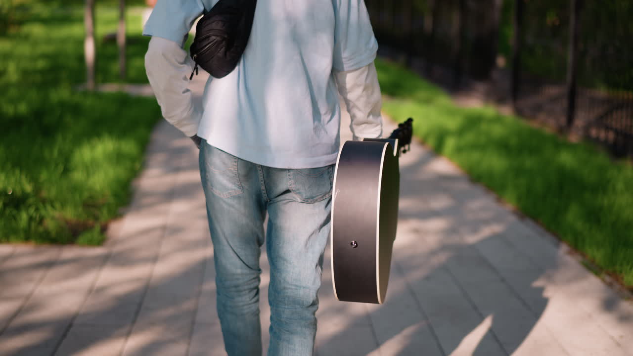 日陰の小道をギターケースとショルダーバッグを持って歩く白人ギタリスト、長い影に対する後姿のシルエット、夕方のギグに向かう、物憂げで内省的な舗装のシーン