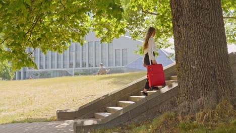 woman caries red suitcase on stairs underneath tree in city center, sunny day