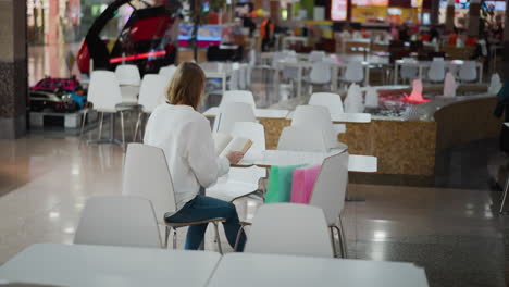 mujer sentada en una mesa en un centro comercial, centrada en leer su libro, con bolsas de compras de colores a su lado, la iluminación suave ilumina el espacio, y una fuente de agua con luces iluminadas es visible