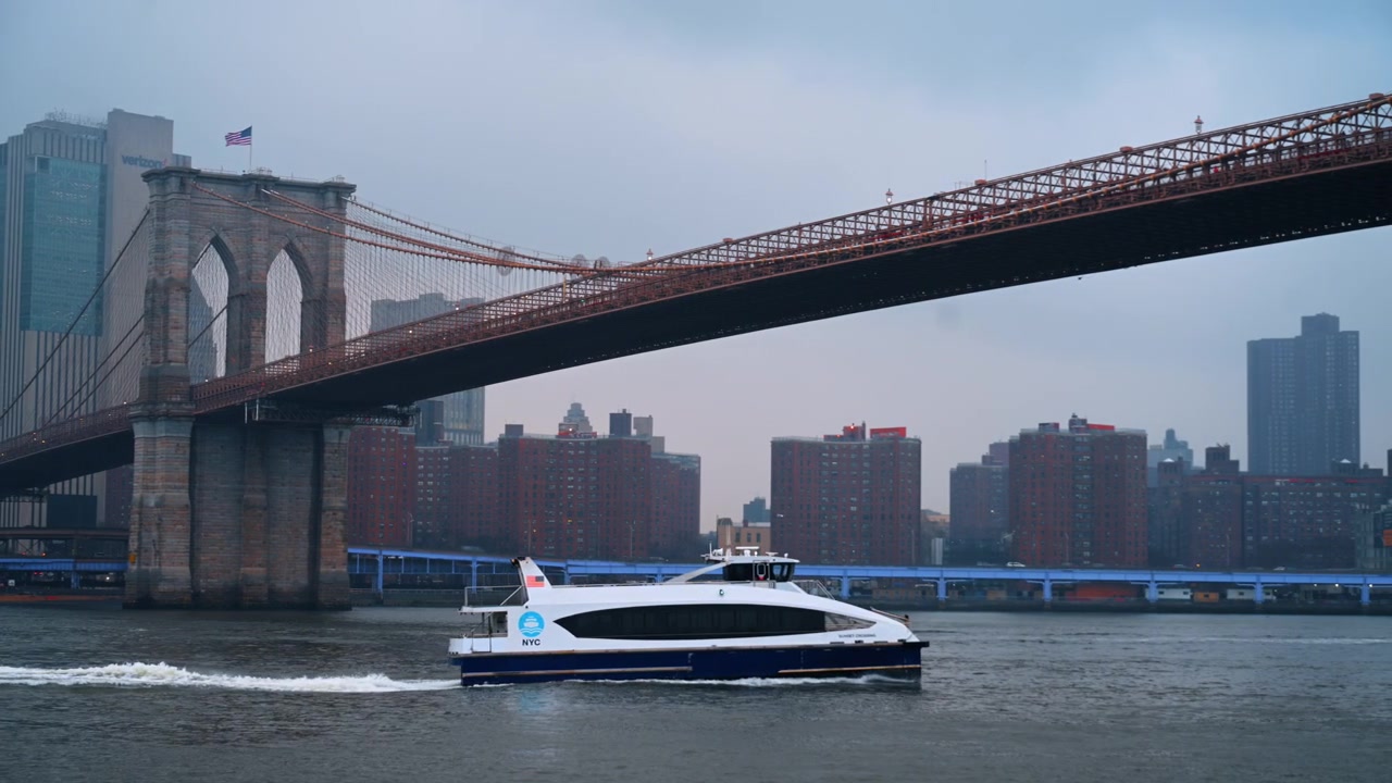 New York, USA, 25 dicembre 2025: Il traghetto NYC passa sotto il ponte di Brooklyn a Manhattan. Il traghetto NYC passa sotto il ponte di Brooklyn sull'East River, lungomare di Manhattan nella foschia, luce blu del crepuscolo.