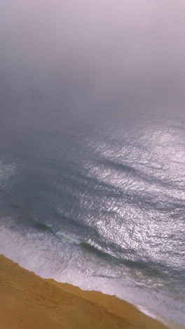 sandy beach with waves moving to it. drone footage over the coastline and ocean waters. thick fog covering the horizon. vertical video