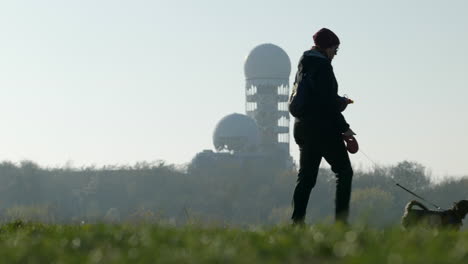 Woman-walking-dog-at-park-with-abandoned-antennas-on-background,-Berlin,-day