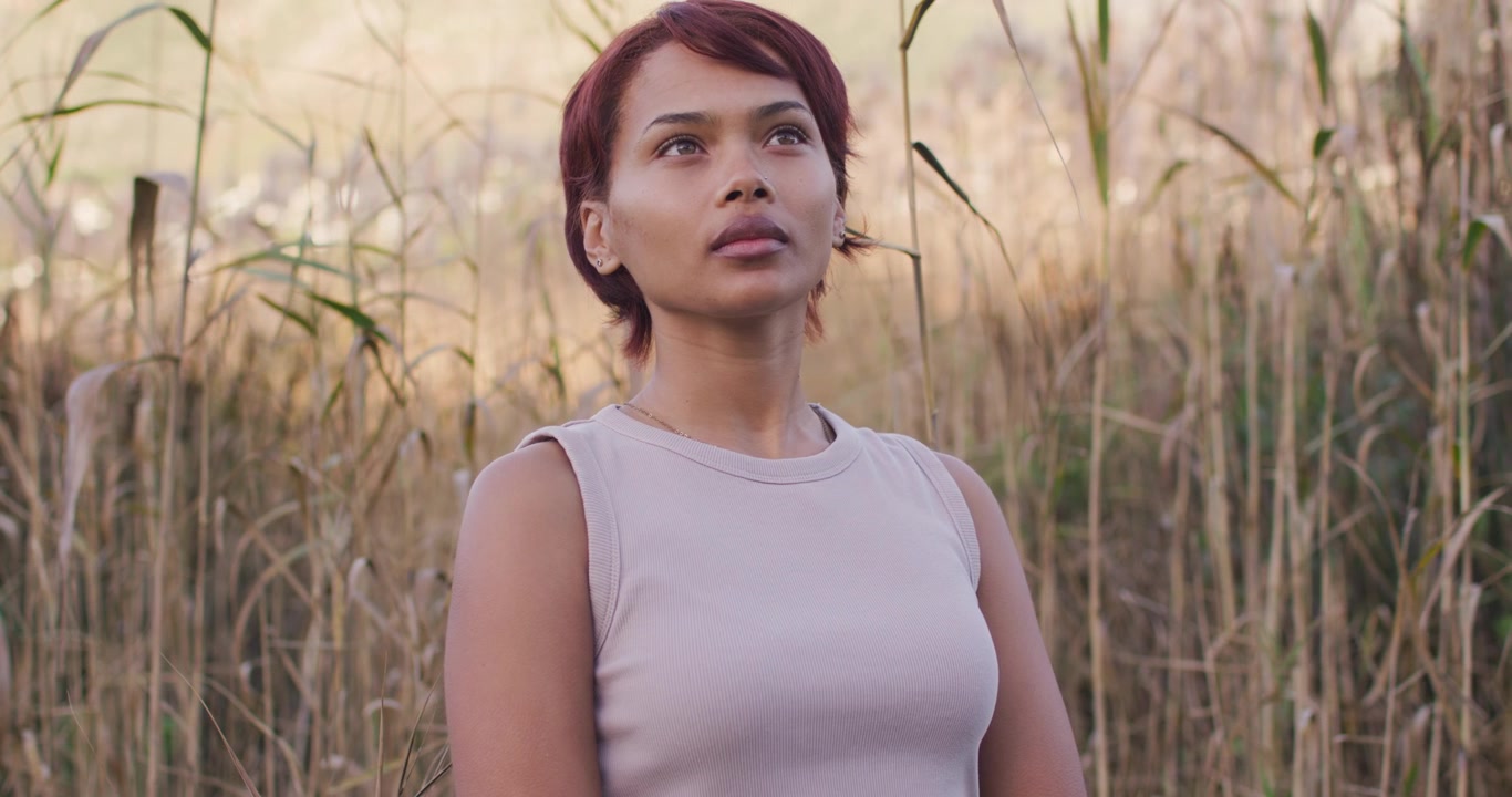 African American woman facing camera, standing in dry grasses, shifting gaze, tilting head, smiling